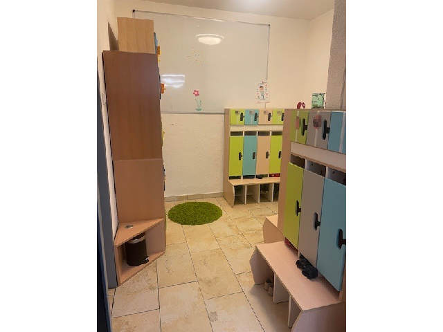 Die Garderobe der Mondgruppe in der Kinderkrippe Sonnenschein. Links an der Wand steht ein Schrank. Rechts befindet sich eine Kindergarderobe mit Sitzfläche und einzelnen Fächern für jedes Kind in grün, blau und grau. Im Hintergrund hängt eine weiße Tafel an der Wand. Auf dem gefliesten Boden liegt ein grüner, runder Teppich.