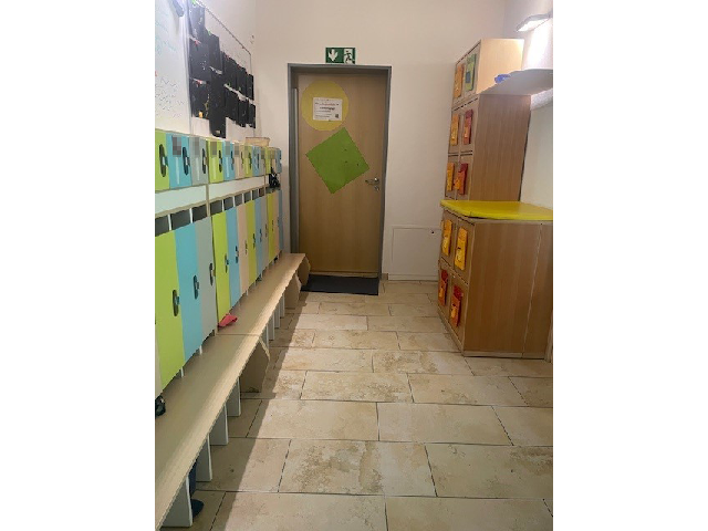 Die Garderobe der Sternengruppe in der Kinderkrippe Sonnenschein. Links an der Wand befindet sich eine Kindergarderobe mit Sitzfläche und einzelnen Fächern für jedes Kind in grün, blau und grau. Rechts an der Wand steht ein Schrank. Im Hintergrund ist eine Tür.