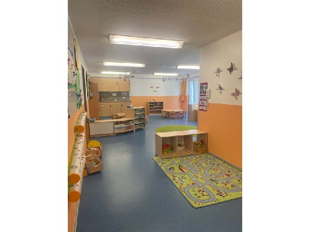 Der Gruppenraum der Mondgruppe in der Kinderkrippe Sonnenschein. Rechts auf dem Boden ist großer Spielteppich und ein Regal mit Spielzeug. Im Hintergrund befindet sich eine Küchenzeile sowie mehrere Tische und Stühle.