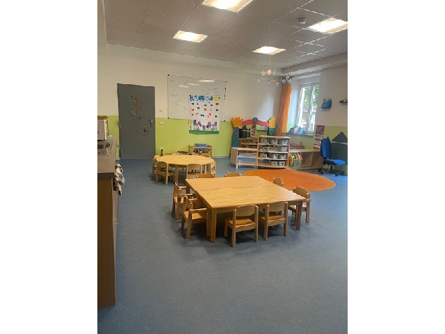 Der Gruppenraum der Sternengruppe in der Kinderkrippe Sonnenschein. Links befindet sich eine Küchenzeile. Im Raum verteilt stehen mehrere Tische und Stühle. Rechts auf dem Boden liegt ein runder, oranger Teppich. Im Hintergrund befindet sich ein Regal mit Spielzeugen, eine weiße Magnettafel sowie eine Tür.