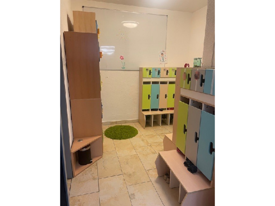 Die Garderobe der Mondgruppe in der Kinderkrippe Sonnenschein. Links an der Wand steht ein Schrank. Rechts befindet sich eine Kindergarderobe mit Sitzfläche und einzelnen Fächern für jedes Kind in grün, blau und grau. Im Hintergrund hängt eine weiße Tafel an der Wand. Auf dem gefliesten Boden liegt ein grüner, runder Teppich.