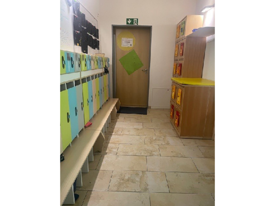 Die Garderobe der Sternengruppe in der Kinderkrippe Sonnenschein. Links an der Wand befindet sich eine Kindergarderobe mit Sitzfläche und einzelnen Fächern für jedes Kind in grün, blau und grau. Rechts an der Wand steht ein Schrank. Im Hintergrund ist eine Tür.