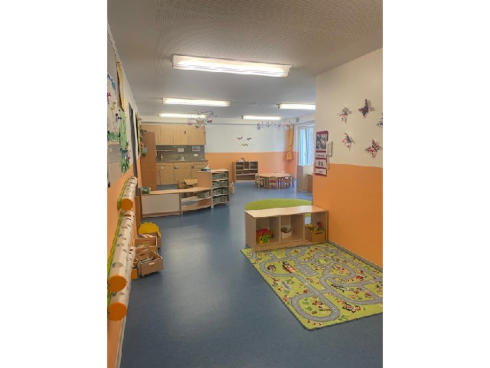 Der Gruppenraum der Mondgruppe in der Kinderkrippe Sonnenschein. Rechts auf dem Boden ist großer Spielteppich und ein Regal mit Spielzeug. Im Hintergrund befindet sich eine Küchenzeile sowie mehrere Tische und Stühle.