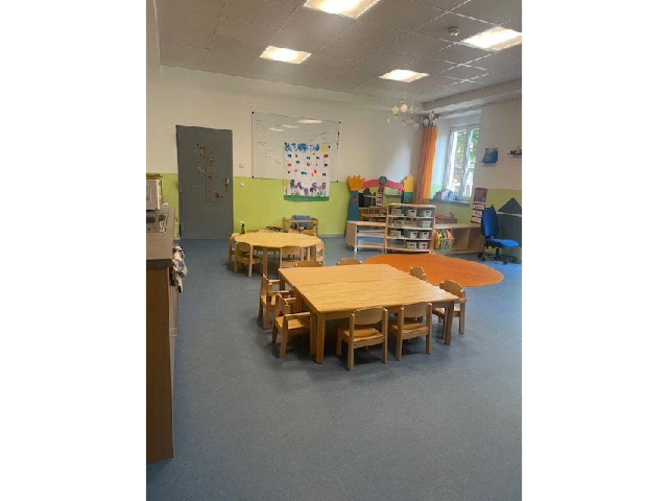 Der Gruppenraum der Sternengruppe in der Kinderkrippe Sonnenschein. Links befindet sich eine Küchenzeile. Im Raum verteilt stehen mehrere Tische und Stühle. Rechts auf dem Boden liegt ein runder, oranger Teppich. Im Hintergrund befindet sich ein Regal mit Spielzeugen, eine weiße Magnettafel sowie eine Tür.