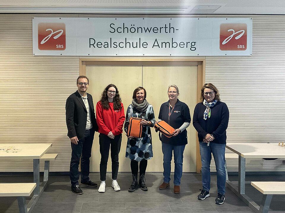 Fünf Personen stehen mit zwei neuen Sanitätstaschen vor einer weißen Banner mit der Aufschrift "Schönwerth-Realschule Amberg". Links und rechts daneben sind Bänke und Tische.