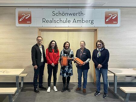 Fünf Personen stehen mit zwei neuen Sanitätstaschen vor einer weißen Banner mit der Aufschrift "Schönwerth-Realschule Amberg". Links und rechts daneben sind Bänke und Tische.
